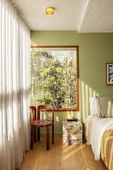 Bedroom with green walls, light hardwood floors, granite stone cube night stand, and wood stool chair with round, dark brown leather cushion, white curtains, and large windows with wooden frames looking out over forest in prefab home in Harpswell, Maine, renovated by Home Union midcentury furniture store in Brooklyn.