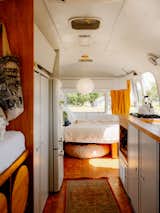 Interior inside of white Argosy airstream from 1975 renovated by Caroline Burke and Riley Haakon with wood tile flooring, oriental rug, globe round spherical pendant light, kitchen with wood countertop and white kettle, and sleeping nook with cream checkered bedspread and yellow curtains.