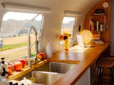 Kitchenette in white Argosy airstream from 1975 renovated by Caroline Burke and Riley Haakon with wood countertop, undermounted sink, and brown leather bar chairs.