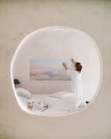 Seen through a round hole in the wall made of chaux lime plaster, a woman hangs painting of clouds in a bedroom with white walls and a bed with a white coverlet as two cats sleep on the bed in a 1000-year-old stone building in Provence, France, renovated by Belgian artist Eva Claessens and converted into her home and studio.