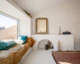 A sculpture studio with a white chaux lime plaster floor, white walls, white ceiling, pink red rug, day bed built into nook beside a long window with an orange brown net coverlet and green pillows, an antique mirror and candlestick, and abstract sculptures arrayed on floor in a 1000-year-old stone building in Provence, France, renovated by Belgian artist Eva Claessens and converted into her home and studio.