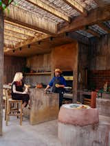 Man behind a concrete bar counter talks to woman sitting on tall light wood bar chair in a concrete addition with a straw roof beside an 1800s brick structure in Todos Santos, Baja California, Mexico, renovated and expanded by Ernesto Kut Gomez, Ellen Odegaard, and Yashar Yektajo.