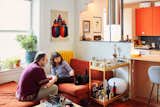 Nicholas Hussong, Masha Tsimring, and their black cat sit on red corduroy sofa chair and ottoman beside yellow bar cart in their Queens, New York, apartment whose kitchen was renovated by architects Lane Rick and Can Vu Bui.