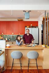 Nicholas Hussong and Masha Tsimring stand in the kitchen of their Queens, New York, with bright orange cabinets turquoise subway tile backsplash, tall chairs with fuzzy grey upholstery, and a wood millwork kitchen island renovated by architects Lane Rick and Can Vu Bui.