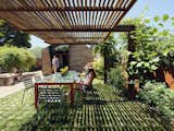 Woman sits beneath wood-slatted shading roof awning at long red table on grassy lawn backyard of accessory dwelling unit ADU in  St. Helena, California CA designed by Ryan and Maddie Chandler of Chandler Workshop Architects.