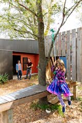 Girl swings on rope in garden beside detached workshop with grey and bright orange metal siding and home in the Black Pearl neighborhood of New Orleans, Louisiana LA, designed by Emilie Taylor and Seth Welty of design firm Colectivo.