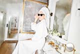 Designer Analisse Taft-Gersten wears white robes and sunglasses as she sits on marble counter of bathroom with medium toned wood flooring.