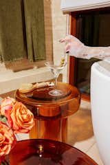 Woman in freestanding tub pulls skewer with olives out of martini glass that sits on red, translucent transparent red orange side table in bathroom with light wood floors and olive green curtain.