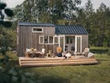 Man and woman recline on long, light wood deck outside Norske Mikrohus tiny house micro house heim adu accessory dwelling unit prefab prefabricated with a metal roof and dark cladding in a forest in Norway.