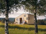 Women friends recline on long, light wood deck outside Norske Mikrohus tiny house micro house tind adu accessory dwelling unit prefab prefabricated with a metal roof and light cladding in a forest glade in Norway.