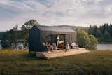 Man and woman recline on long, light wood deck as man stands in doorway of Norske Mikrohus tiny house micro house rast adu accessory dwelling unit prefab prefabricated with a metal roof and dark cladding in a forest in Norway by a river.