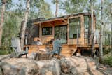 Fritz Tiny Home Halcyon model with metal and wood siding and wraparound porch sits in a clearing in a forest with a bonfire circled by outdoor chairs.