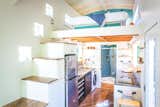 Interior of Oasis Tiny Home by Paradise Tiny Homes in Hawaii with white interior, staircase with dark wood tread that leads up to bedroom loft, glossy kitchen cabinet, metal sink, medium-tone wood floors, exposed curved ceiling beams, and blue tile kitchen backsplashes.
