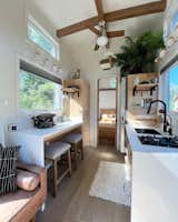Interior of Paradise Tiny Home's Paradise Model with kitchen with white glossy counter tops, tall ceilings with exposed brown beams, white built-in dining table set against wall, medium-toned wood floors, octagonal peekaboo window, and tall doorway leading into master bedroom in Hawaii Hawai'i.