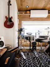 Interior of Noyer prefab tiny mobile home model by Minimaliste with white painted engineered wood walls, pine ceiling, vinyl flooring, large dark rug with geometric line pattern, red guitar hanging from wall, and drum set sitting beneath wide window.