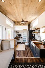 Interior of Noyer prefab tiny mobile home model by Minimaliste with white painted engineered wood walls, pine ceiling, vinyl flooring, blue kitchen cabinetry, dark stained countertops, and work station set up against far wall beneath loft.