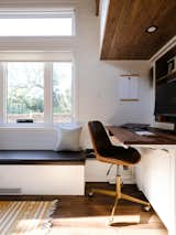 Interior of Noyer prefab tiny mobile home model by Minimaliste with white painted engineered wood walls, vinyl flooring, long white bench with black leather cushion, and workstation with desk with dark-toned wood top and brass rolling desk chair with seat upholstered in brown leather.