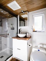 Bethroom of Noyer prefab tiny mobile home model by Minimaliste with white painted engineered wood walls, cedar ceiling, vinyl flooring, white vanity, dark wood counter top, white pedestal bowl sink, and glass enclosed shower booth.