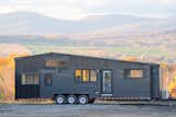 Minimaliste prefab tiny mobile home Noyer XL model with blacked cedar and standing seam metal siding sits parked in on a gravelly road overlooking an autumn landscape with farms and rolling hills.