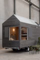 ROAM tiny mobile home on a three-axle trailer by Fabprefab with grey Accoya timber lap siding and corner wrap around windows. Outdoor green metal patio furniture and and leafy plant sit outside it.