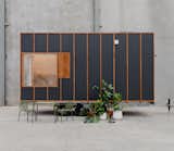 ROAM tiny mobile home on a three-axle trailer by Fabprefab with blackened Weathertex siding set in timber framing, and corner wrap around windows. Outdoor green metal patio furniture and and leafy plants in white and terracotta pots sit outside it.