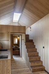 Interior of tiny home by Madeiguincho with walls clad in pale wood, pitched ceiling with long skylight with shutter, a rail-less staircase with medium-toned wood tread leading up to a lofted bedroom, combined kitchen and hallway, and bathroom at the end of the hallway.