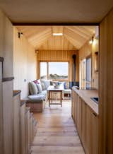 Combined living room and kitchen in tiny home by Madeiguincho with with wood flooring, medium-toned wood walls, wood cabinets, wraparound sofa bench in corner of unit, and large window with view of sandy beach.
