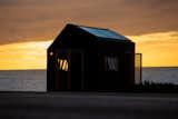 Tiny home by Madeiguincho with blackened cladding, large skylights, and wood slated shutters sits on a trailer on beach.