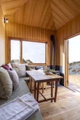 Living room of tiny home by Madeiguincho with medium-toned wood walls, wood-burning stove, wraparound sofa bench built into wall, a tall coffee table, and large doors and window with views of sandy beach.