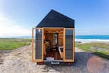 View of interior of tiny home by Madeiguincho with blackened cladding sitting on a trailer on rocky beach through open tall glass doors at the end of unit.