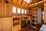 Kitchen of tiny home by Madeiguincho with unfinished wood framed walls, large skylights, wood counter, open wooden cabinetry, exposed wires and utilities, dark wood floors, and wooden ladder leading up to lofted bedroom.
