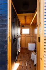 Bathroom of tiny home by Madeiguincho with unfinished wood framed walls, black wall with black showerhead, and white one-piece toilet.