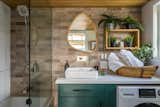 Bathroom of Summit Tiny Homes Nysos model with dark green vanity, white Corian countertop, pedestal sink, curving asymmetric mirror, steel fixtures, brick veneer accent wall, oak shelf with plants, and combined washer dryer machine.