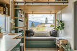 Interior of Summit Tiny Homes Nysos model with ladder leading up to lofted sleeping area bedroom and reading nook with slatted oak ceiling, dark green cabinetry, dark green library book shelves, and dark green sofa beside window with cushion upholstered in grey fabric.