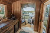 The interior of Vagabond Haven's prefab tiny called Nature Pod with pine floors and walls, Thermowood kitchen cabinetry, living room bedroom with panoramic window, blue-grey upholstered chair and futon and yellow rug.