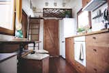 Interior of Vagabond Haven's prefab tiny home called Elise with white painted spruce walls, dark wood veneer laminate floors, MDF kitchen cabinetry, stowable fold-out tables with box stools, and ladder leading up to lofted bedroom.