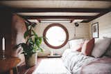 Den with round window, mattress, plant in whicker pot, wall lamp, end table, white rug, and MDF storage cubby beneath raised living room in Vagabond Haven's prefab tiny home called Elise.