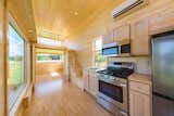 Interior of ESCAPE homes prefab tiny home ONE model with staircase leading up to loft with white metal railings, birch floors, birch walls, birch ceiling, and light wood cabinetry in kitchenette with oven, stove, overhead microwave, and sink.