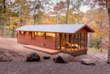 ESCAPE homes prefab tiny home classic model with red cedar cladding, deck, roofed and enclosed patio, and metal roof sits on shore of pond in autumn forest.