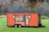 ESCAPE homes prefab tiny home vista boho model with red vertical wood cladding sits on a trailer in a grassy field.