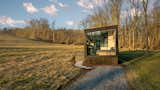 Zook Cabins and New Frontiers Design made the Luna Park Model Tiny Home with blackened metal and cedar cladding and floor-to-ceiling glass window at the end of gravel driveway in a field.