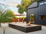 Four people talk beneath steel pergola in gravel backyard with gabion L-shaped bench, firepit, and corrugated metal fencing designed by designed by Ryan McWhirter of Lush GreenScape Design outside architect Karin Scott and Don Doughtery’s gabled, black metal-clad home in Terrell Hills neighborhood of San Antonio, Texas TX.