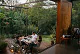 Three people sit outside brick and gravel patio terrace outside extension of home in Nairobi, Kenya, designed by Studio Propolis, cofounded by Naeem Biviji and Benthan Rayner, with long red-brown table, white wire frame chairs, metal-framed canopy roof with wire stretchers, and planters with shrubs.