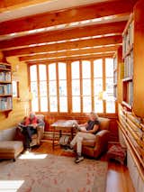 Man and woman sit reading in brown leather armchairs in library in schooner docked in Point Harbor in Sausalito, CA, renovated by Ryan Phelan and Stewart Brand, with end table, large red, beige, and green rug with floral designs, medium-toned wood floors and walls, tall casement windows, swiveling brass wall lamps, and exposed wood ceilings.