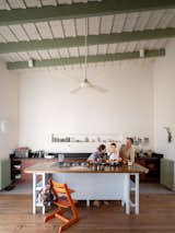 Man, woman, and boy sit at kitchen island with dark wood top, white grey legs and base in kitchen with medium-toned wood floors, red-brown cabinetry, black countertops, light olive green radiators and exposed ceiling beams, tall ceilings, and shelf recessed into wall in home in the old quarter of San Antonio de Areco, Argentina, renovated by Julián Benedit Prebisch and Micaela Suide.