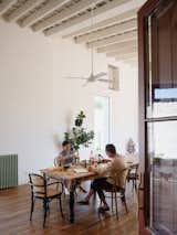Man and woman eat at long table with dark legs, flaking paint, and light wood counter top, black chairs with rattan seats and backing, and tall ceilings with exposed beams in home in the old quarter of San Antonio de Areco, Argentina, renovated by Julián Benedit Prebisch and Micaela Suide.