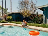 Woman sits on coping beside dog while she reads a book and dips legs in pool with orange tile band beneath coping designed by Jared Frank Studio and built by Blue Pacific Contractors outside Los Angeles home.