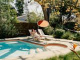 Woman reads a book on white lawn chair beneath bright orange parasol beside pool with orange tile band beneath coping designed by Jared Frank Studio and built by Blue Pacific Contractors outside Los Angeles home.