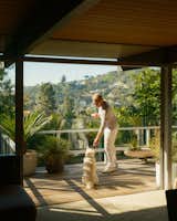 Patrick Thomas O’Neill plays with white dog on porch of the Hailey House by Richard Neutra renovated by Anthony Barsoumian overlooking garden designed by Studio John Sharp with white metal frame and glass railing, wood decking, and potted plants.