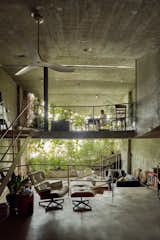 Man works at desk in top floor office overlooking living room with Eames chair upholstered in white leather in split-level Singapore home by Linghao architects with concrete floors, wall, and ceiling, and open windows looking out into verdant overgrown garden.